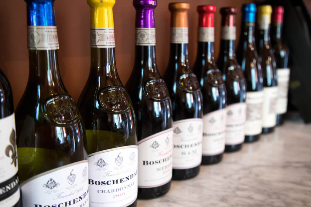 A row of wine bottles with rainbow colored tops. Visiting the Boschendal Wine Farm in South Africa is a treat for the senses. From delectable food to beautiful scenery to the savory wine, it's an experience that shouldn't be missed in South Africa. Here's what you need to know before you go. And for more travel tips and wanderlust inspiration, follow The Scenic Suitcase on Pinterest!