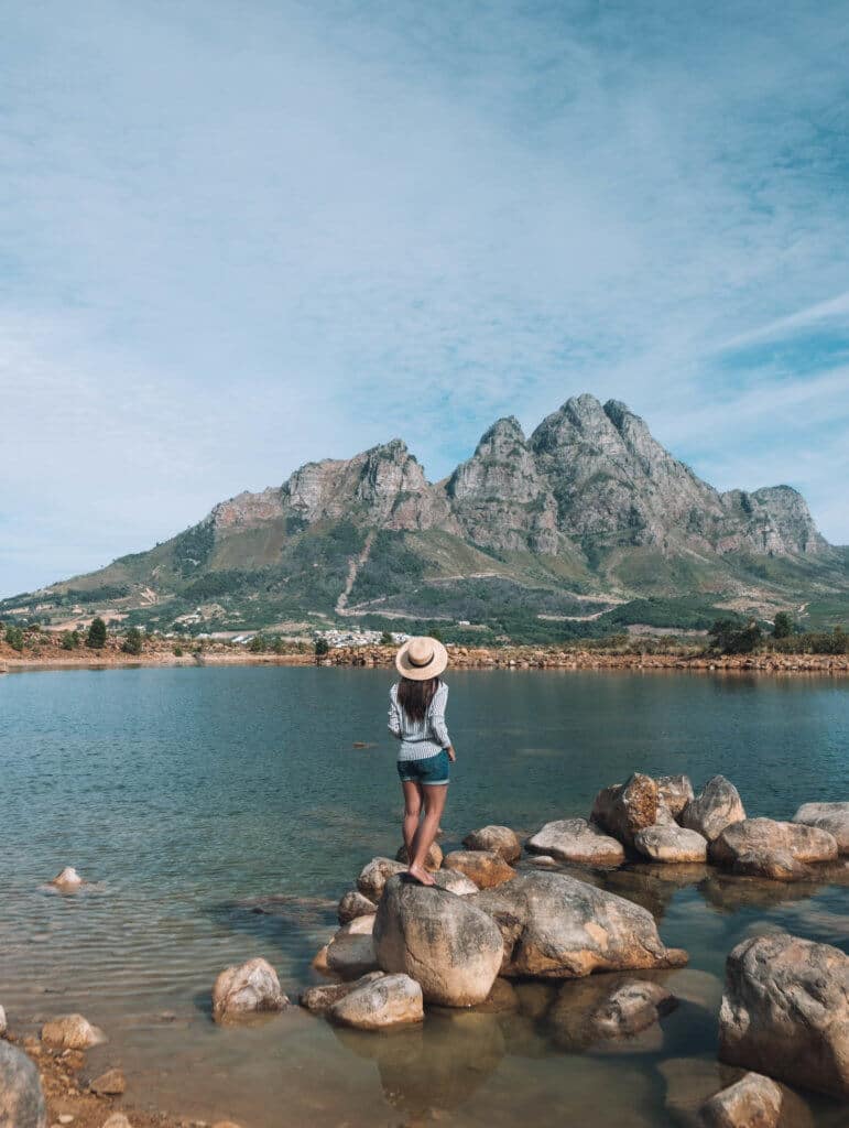 Girl standing on a rock at the edge of a lake looking towards the mountains. Visiting the Boschendal Wine Farm in South Africa is a treat for the senses. From delectable food to beautiful scenery to the savory wine, it's an experience that shouldn't be missed in South Africa. Here's what you need to know before you go. And for more travel tips and wanderlust inspiration, follow The Scenic Suitcase on Pinterest!