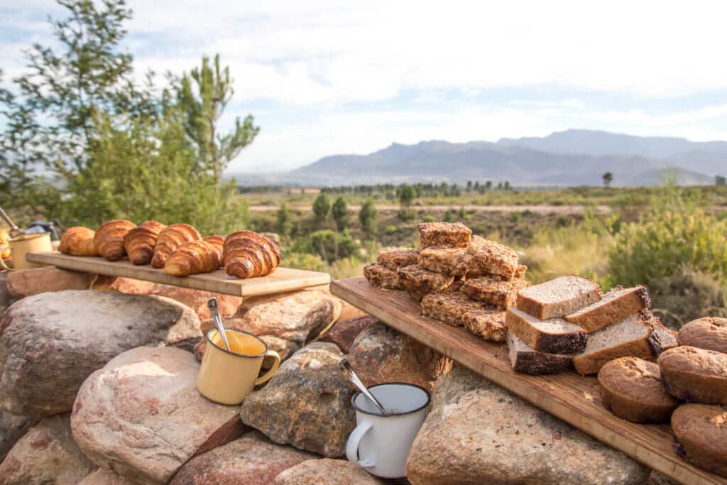 A variety of breakfast breads balanced on rocks with the mountains in the background. Visiting the Boschendal Wine Farm in South Africa is a treat for the senses. From delectable food to beautiful scenery to the savory wine, it's an experience that shouldn't be missed in South Africa. Here's what you need to know before you go. And for more travel tips and wanderlust inspiration, follow The Scenic Suitcase on Pinterest!