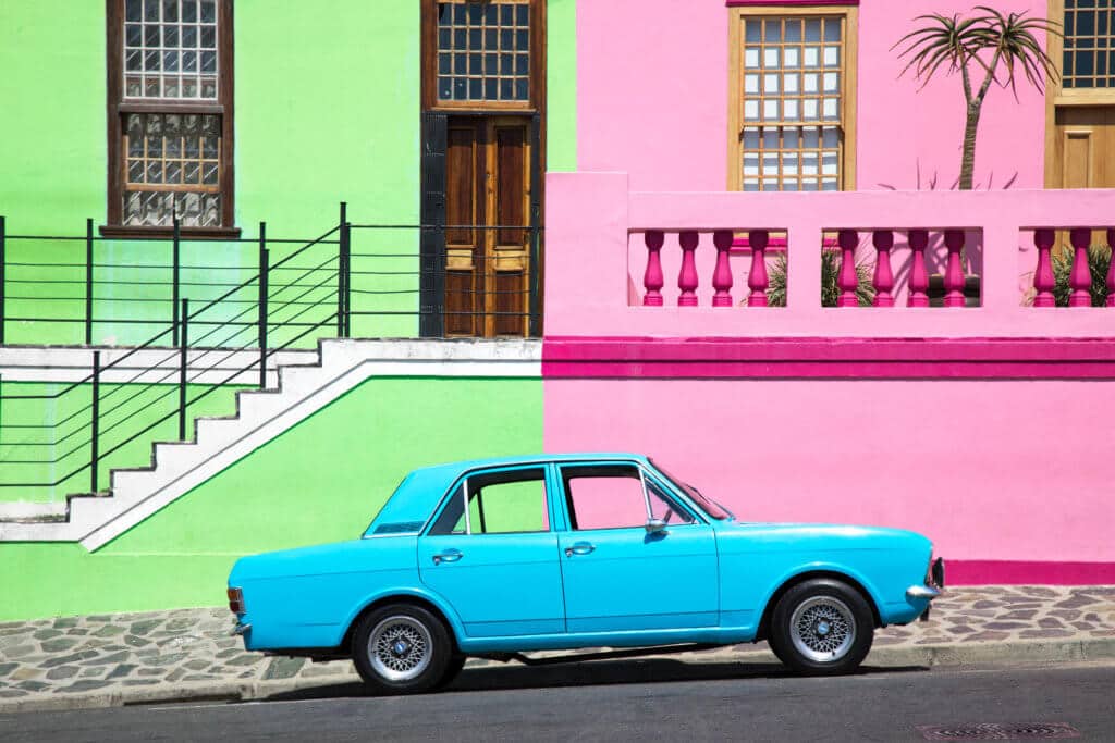 A green and pink building with a blue car parked in the middle out front. Visiting Bo Kaap in Cape Town feels like stepping into a cartoon. But there's more to the neighborhood than prismatic facades. Discover the colorful history of one of South Africa's most intriguing communities. Plus, get expert tips on what you need to know before you go! And for more travel tips, follow The Scenic Suitcase on Pinterest!