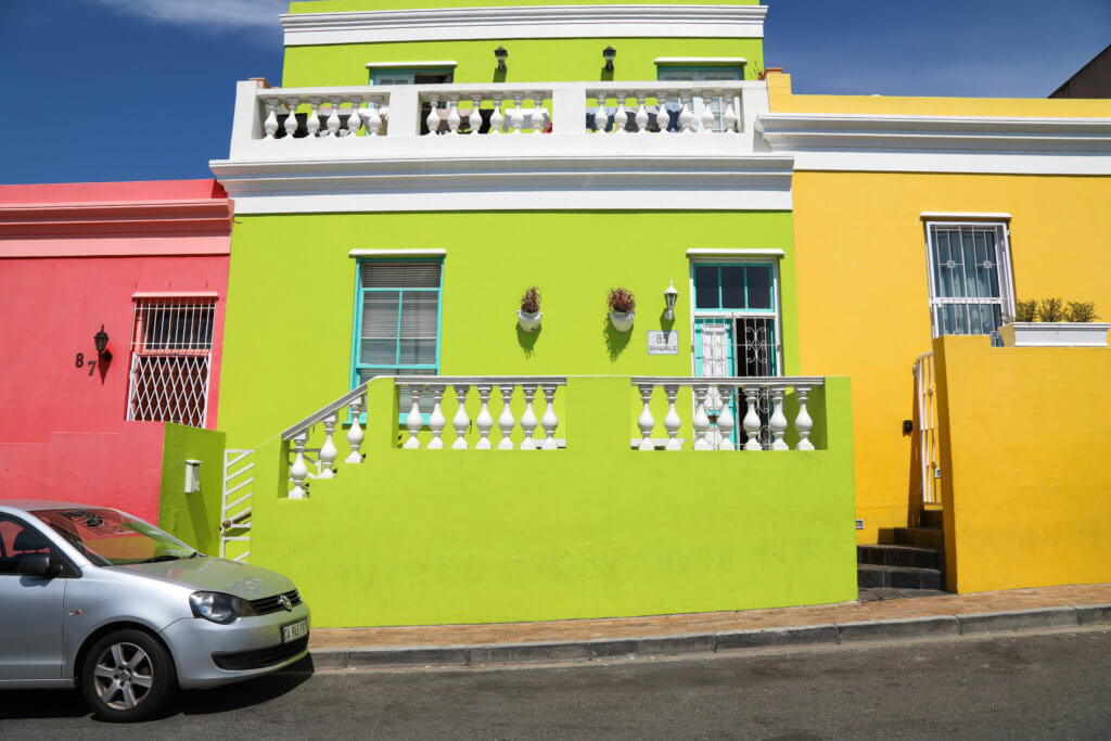 A pink, green, and yellow exterior with a silver car parked out front. Visiting Bo Kaap in Cape Town feels like stepping into a cartoon. But there's more to the neighborhood than prismatic facades. Discover the colorful history of one of South Africa's most intriguing communities. Plus, get expert tips on what you need to know before you go! And for more travel tips, follow The Scenic Suitcase on Pinterest!