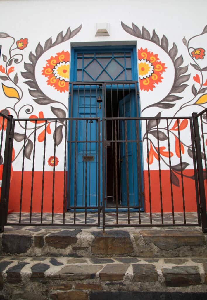 A blue doorway with an orange flower ornately painted around it. Visiting Bo Kaap in Cape Town feels like stepping into a cartoon. But there's more to the neighborhood than prismatic facades. Discover the colorful history of one of South Africa's most intriguing communities. Plus, get expert tips on what you need to know before you go! And for more travel tips, follow The Scenic Suitcase on Pinterest!
