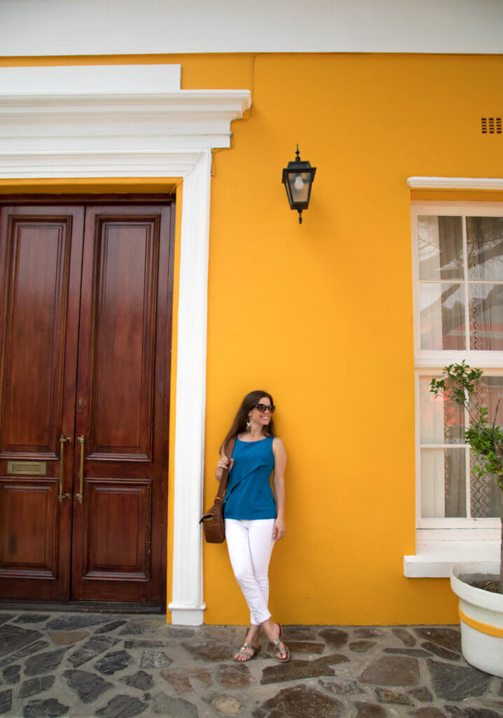 A girl dressed in blue and white standing next to a bright yellow building. Visiting Bo Kaap in Cape Town feels like stepping into a cartoon. But there's more to the neighborhood than prismatic facades. Discover the colorful history of one of South Africa's most intriguing communities. Plus, get expert tips on what you need to know before you go! And for more travel tips, follow The Scenic Suitcase on Pinterest!