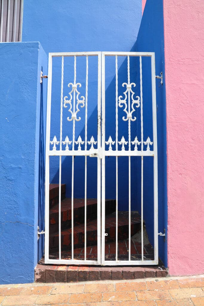 A white gate on a blue and pink building. Visiting Bo Kaap in Cape Town feels like stepping into a cartoon. But there's more to the neighborhood than prismatic facades. Discover the colorful history of one of South Africa's most intriguing communities. Plus, get expert tips on what you need to know before you go! And for more travel tips, follow The Scenic Suitcase on Pinterest!
