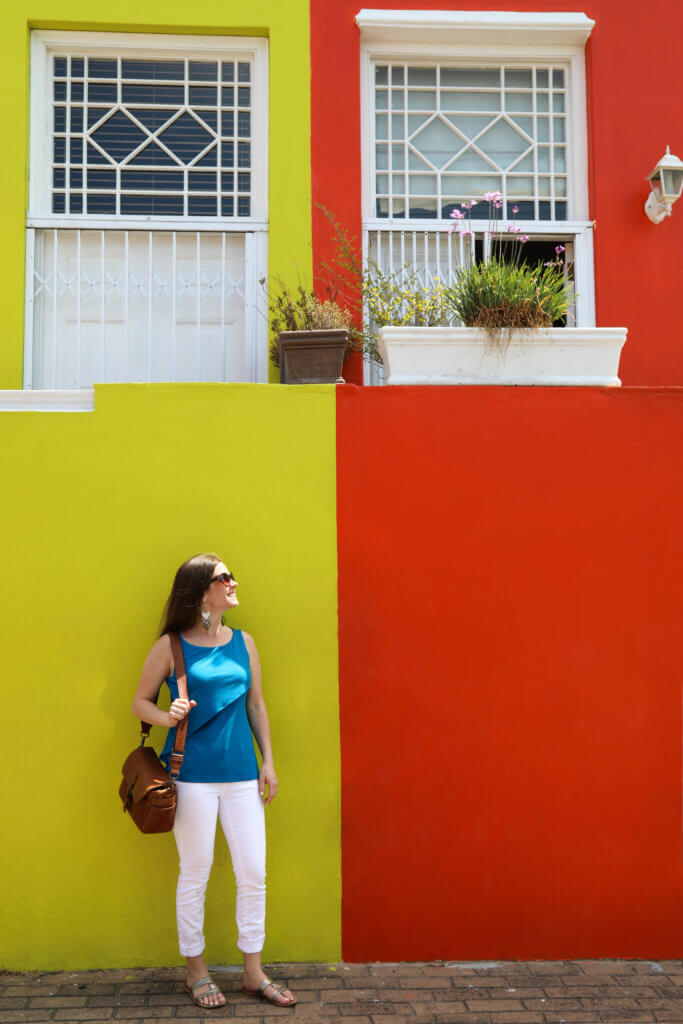 A yellow and orange building behind a girl dressed in white and blue. Visiting Bo Kaap in Cape Town feels like stepping into a cartoon. But there's more to the neighborhood than prismatic facades. Discover the colorful history of one of South Africa's most intriguing communities. Plus, get expert tips on what you need to know before you go! And for more travel tips, follow The Scenic Suitcase on Pinterest!