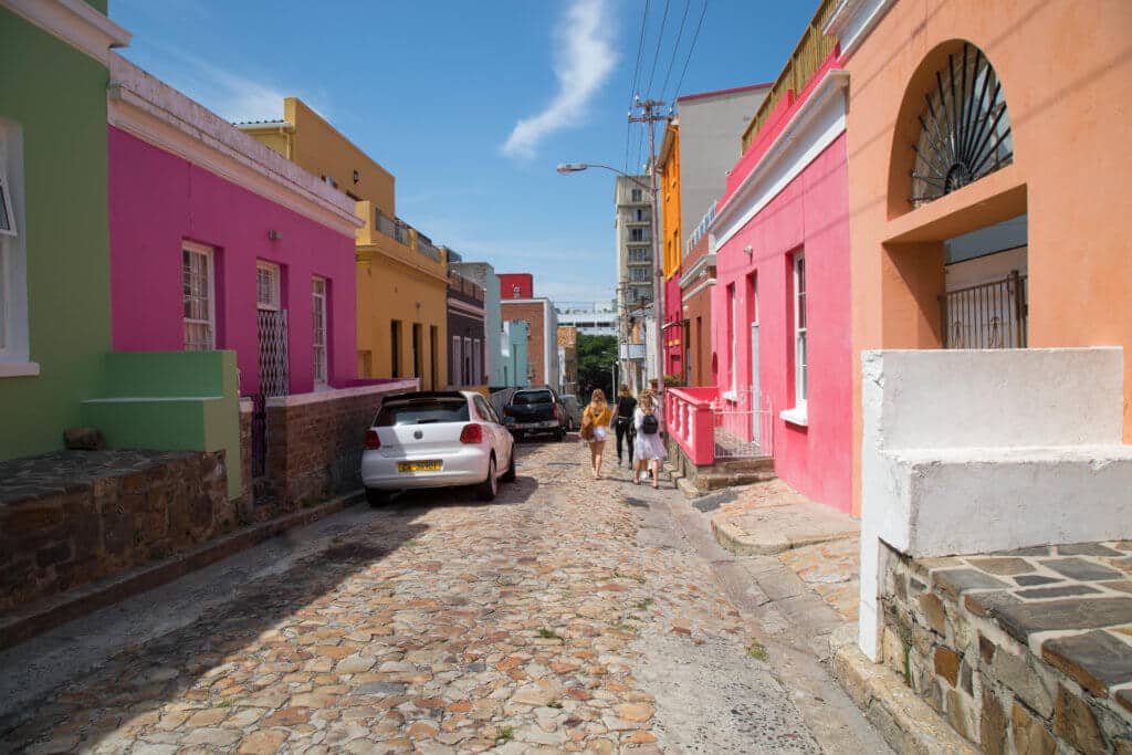 A cobblestone street running through multi-colored buildings. Visiting Bo Kaap in Cape Town feels like stepping into a cartoon. But there's more to the neighborhood than prismatic facades. Discover the colorful history of one of South Africa's most intriguing communities. Plus, get expert tips on what you need to know before you go! And for more travel tips, follow The Scenic Suitcase on Pinterest!
