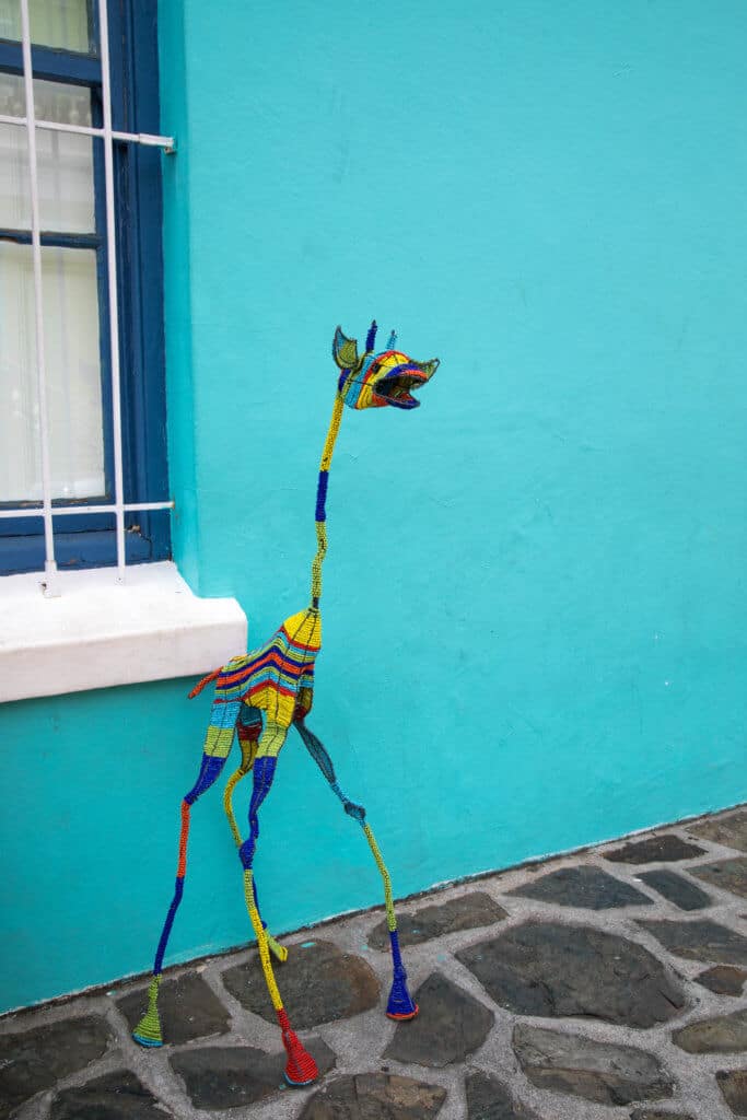 A bead giraffe in front of a blue building window. Visiting Bo Kaap in Cape Town feels like stepping into a cartoon. But there's more to the neighborhood than prismatic facades. Discover the colorful history of one of South Africa's most intriguing communities. Plus, get expert tips on what you need to know before you go! And for more travel tips, follow The Scenic Suitcase on Pinterest!