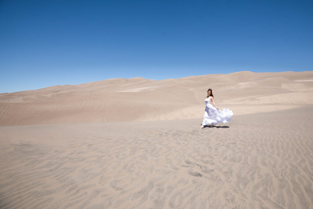Girl in a white dress walking along sand dunes. Get the inside scoop about visiting the Great Sand Dunes from a Colorado local. From where to find them to tips when you get there, this guide has it all! And for more travel tips and wanderlust inspiration, follow The Scenic Suitcase on Pinterest!