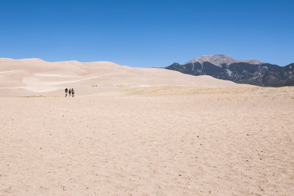 People walking across sand dunes with mountains in the background. Get the inside scoop about visiting the Great Sand Dunes from a Colorado local. From where to find them to tips when you get there, this guide has it all! And for more travel tips and wanderlust inspiration, follow The Scenic Suitcase on Pinterest!