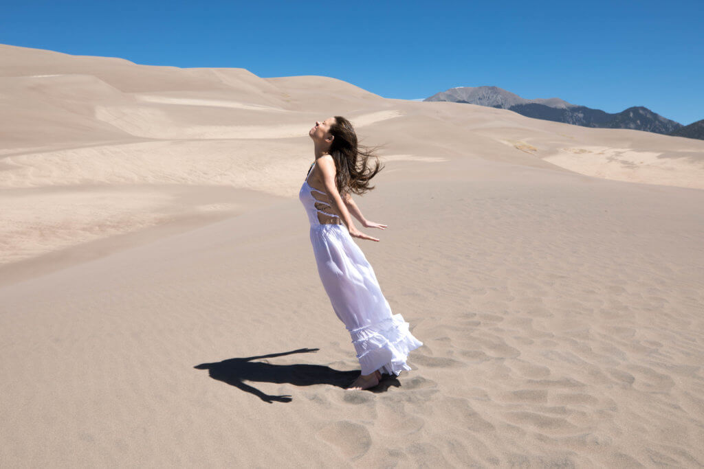 Girl in a white dress being blown away by heavy winds at the sand dunes. Get the inside scoop about visiting the Great Sand Dunes from a Colorado local. From where to find them to tips when you get there, this guide has it all! And for more travel tips and wanderlust inspiration, follow The Scenic Suitcase on Pinterest!