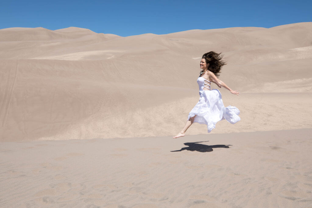 A girl jumping through sand dunes. Get the inside scoop about visiting the Great Sand Dunes from a Colorado local. From where to find them to tips when you get there, this guide has it all! And for more travel tips and wanderlust inspiration, follow The Scenic Suitcase on Pinterest!