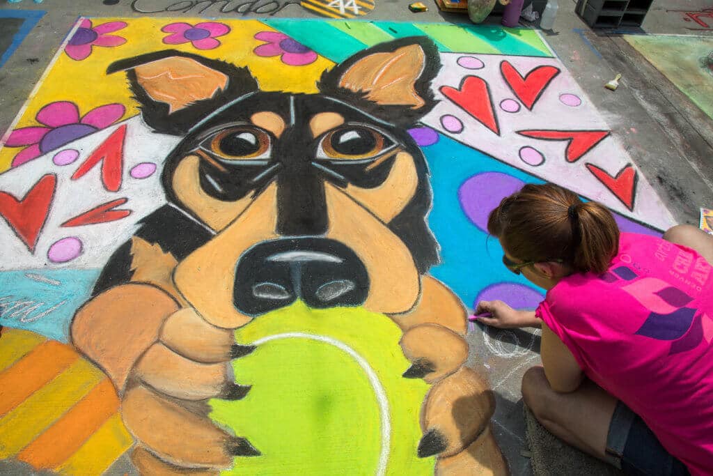 A woman drawing a dog holding a ball with chalk. Find out everything you need to know about the annual Denver Chalk Art Festival! What it's all about, what you can expect, and what you can't miss! And for more travel tips and wanderlust inspiration, follow The Scenic Suitcase on Pinterest!