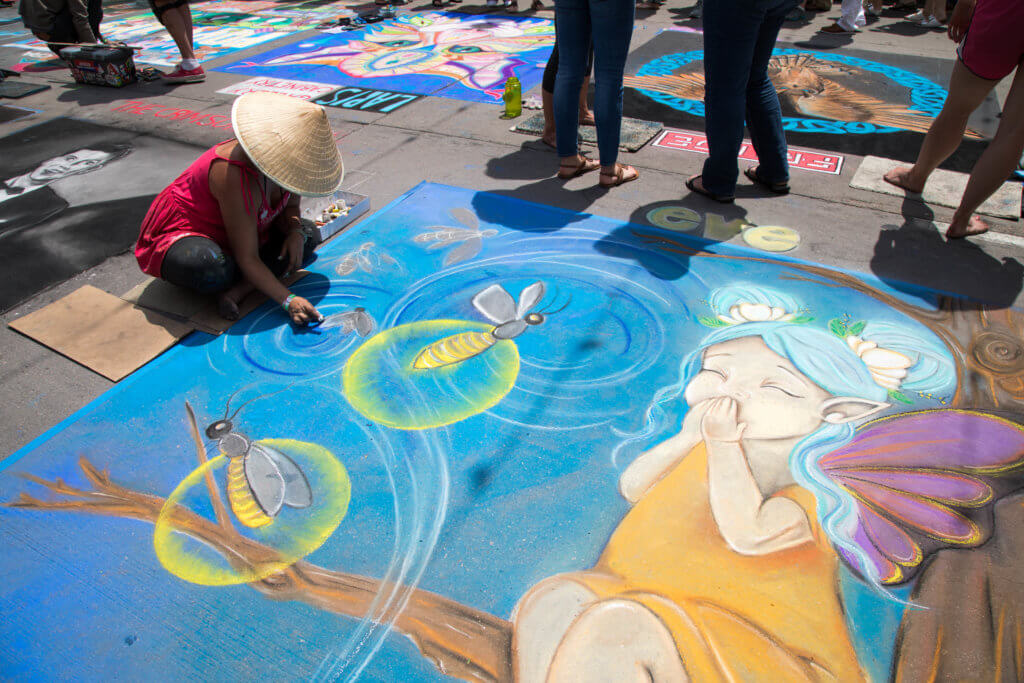 A woman in a hat drawing a giggling fairy in chalk. Find out everything you need to know about the annual Denver Chalk Art Festival! What it's all about, what you can expect, and what you can't miss! And for more travel tips and wanderlust inspiration, follow The Scenic Suitcase on Pinterest!