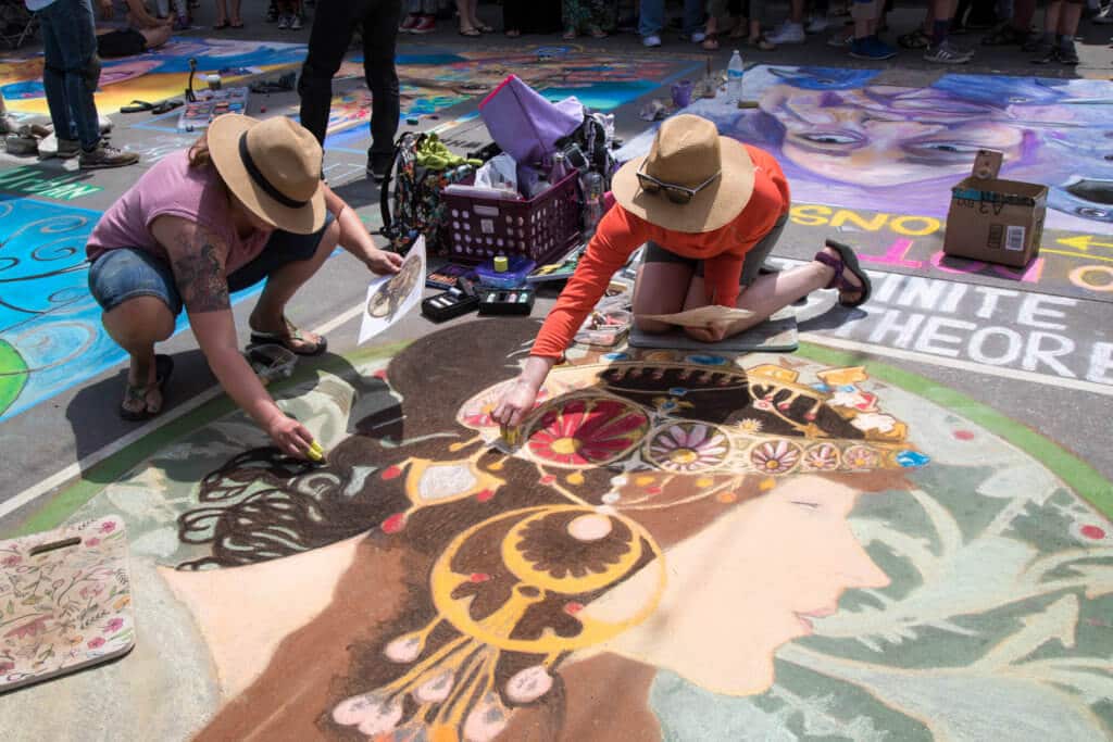 Two women in hats drawing with chalk on the pavement. Find out everything you need to know about the annual Denver Chalk Art Festival! What it's all about, what you can expect, and what you can't miss! And for more travel tips and wanderlust inspiration, follow The Scenic Suitcase on Pinterest!