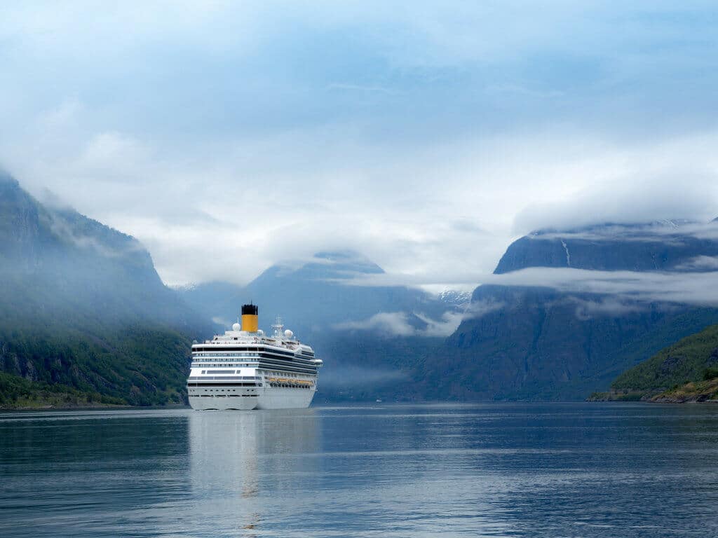 A cruise ship sailing through a fiord surrounded by mountains. Use these secret travel hacks for better flights, hotels, and cruises! Along with tips on how to avoid lines, pick the best travel destinations, and more! And for more travel tips and wanderlust inspiration, follow The Scenic Suitcase on Pinterest!