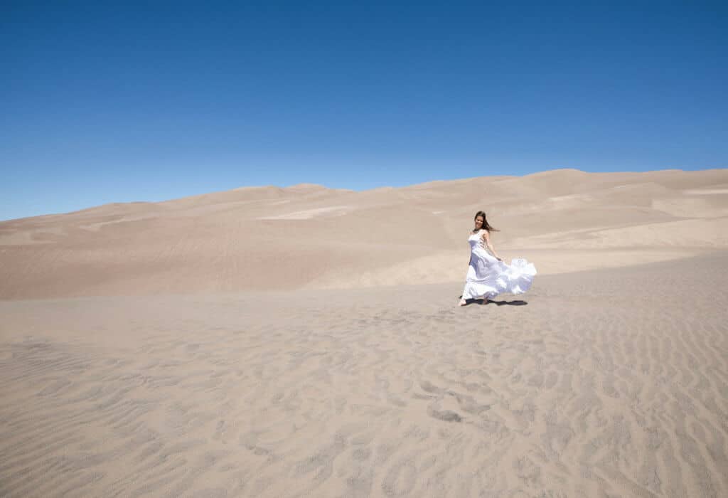 A girl in a white dress walking through sand dunes. Discover the most stunning, offbeat, and unique places in Colorado. Including cliff diving dinner entertainment, haunted hotels, towering dunes, and more! And for additional travel tips and wanderlust inspiration, follow The Scenic Suitcase on Pinterest!