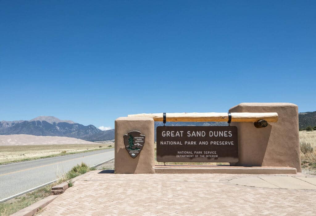 The park entrance sign at the Great Sand Dunes with mountains in the background. Discover the most stunning, offbeat, and unique places in Colorado. Including cliff diving dinner entertainment, haunted hotels, towering dunes, and more! And for additional travel tips and wanderlust inspiration, follow The Scenic Suitcase on Pinterest!