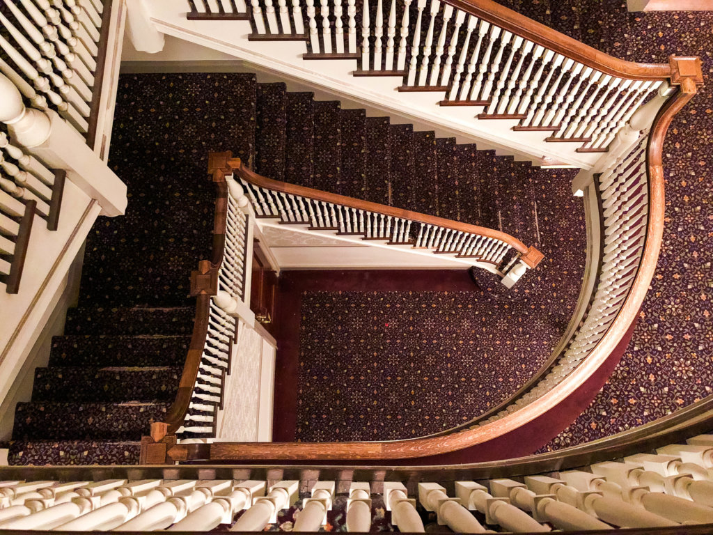 Looking down a stair landing from the top floor of a hotel. The Stanley Hotel has classic charm, an intriguing past, and otherwordly guests. Discover what it's like to meet ghosts on a Stanley Hotel haunted tour! And for more travel tips and wanderlust inspiration, follow The Scenic Suitcase on Pinterest!