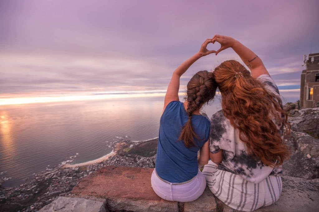 Two girls sitting on a ledge at sunset making a heart with their two hands. See the most picture perfect moments of 2018. From London to Cape Town to Las Vegas and more, see what you can't miss in these incredible destinations! And for more travel tips and wanderlust inspiration, follow The Scenic Suitcase on Pinterest!