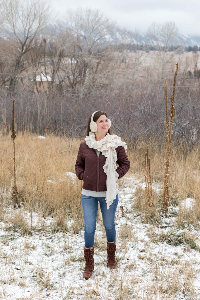 Girl walking through a snowy field in a brown jacket wearing cream ear muffs and a cream scarf. The ultimate winter packing guide with a complete list of items you need for winter travel. Everything you need to stay warm and toasty! And for more travel tips and wanderlust inspiration, follow The Scenic Suitcase on Pinterest!