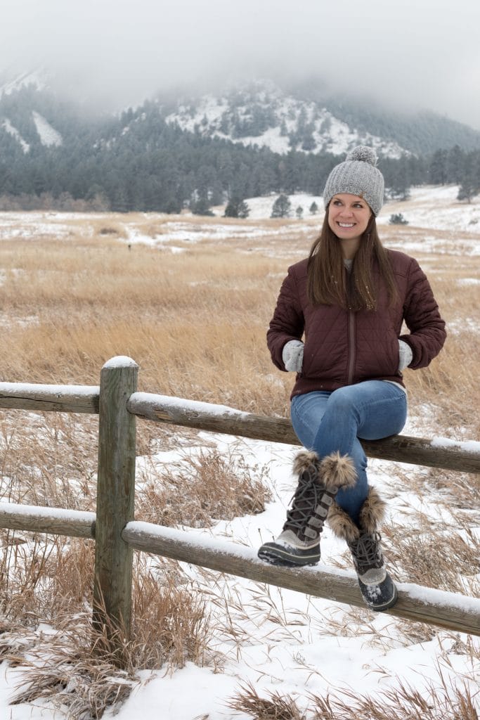 Gir wearing a brown down jacket sitting on a fence post in front of snow mountains. The ultimate winter packing guide with a complete list of items you need for winter travel. Everything you need to stay warm and toasty! And for more travel tips and wanderlust inspiration, follow The Scenic Suitcase on Pinterest!