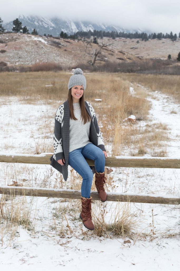 Girl in a long, gray cardigan sittign on a snowy fence post in the mountains. The ultimate winter packing guide with a complete list of items you need for winter travel. Everything you need to stay warm and toasty! And for more travel tips and wanderlust inspiration, follow The Scenic Suitcase on Pinterest!