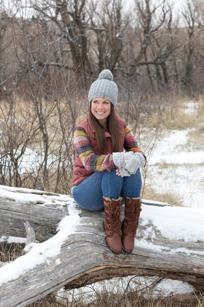 Girl in a pink plaid shirt, pink down vest, jeans, and brown Sorel boots sitting on a snowy log. The ultimate winter packing guide with a complete list of items you need for winter travel. Everything you need to stay warm and toasty! And for more travel tips and wanderlust inspiration, follow The Scenic Suitcase on Pinterest!