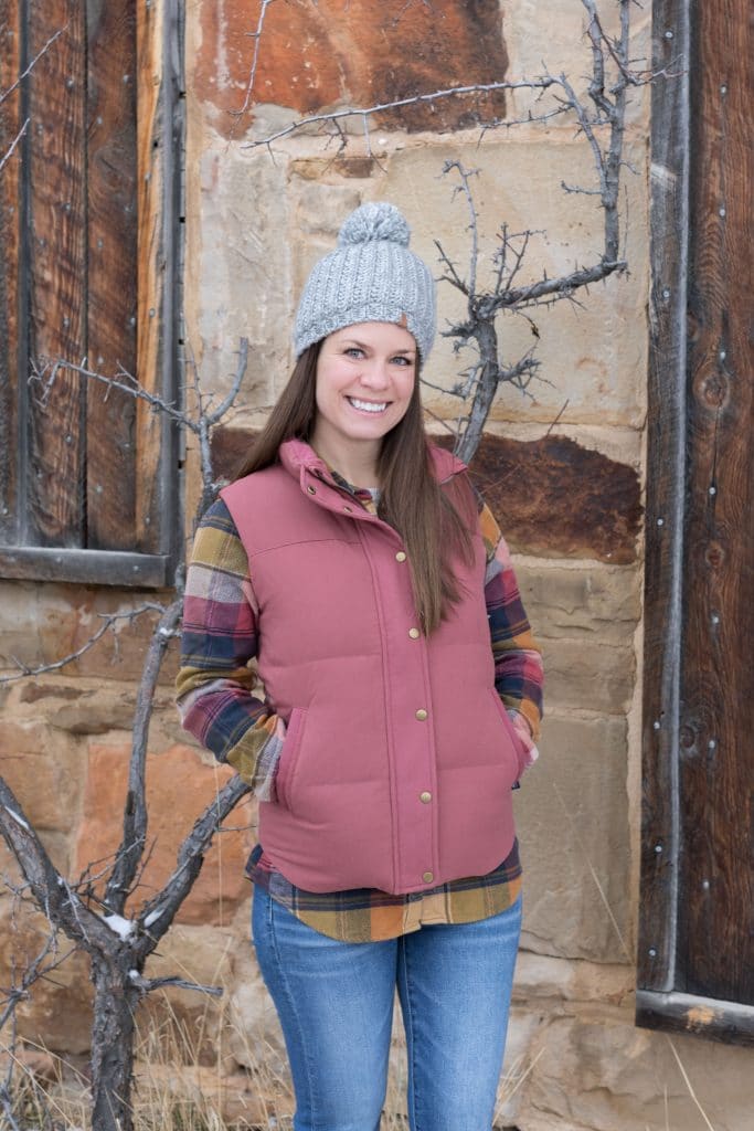 Girl in plaid shirt and pink down vest standing in front of stone building. The ultimate winter packing guide with a complete list of items you need for winter travel. Everything you need to stay warm and toasty! And for more travel tips and wanderlust inspiration, follow The Scenic Suitcase on Pinterest!