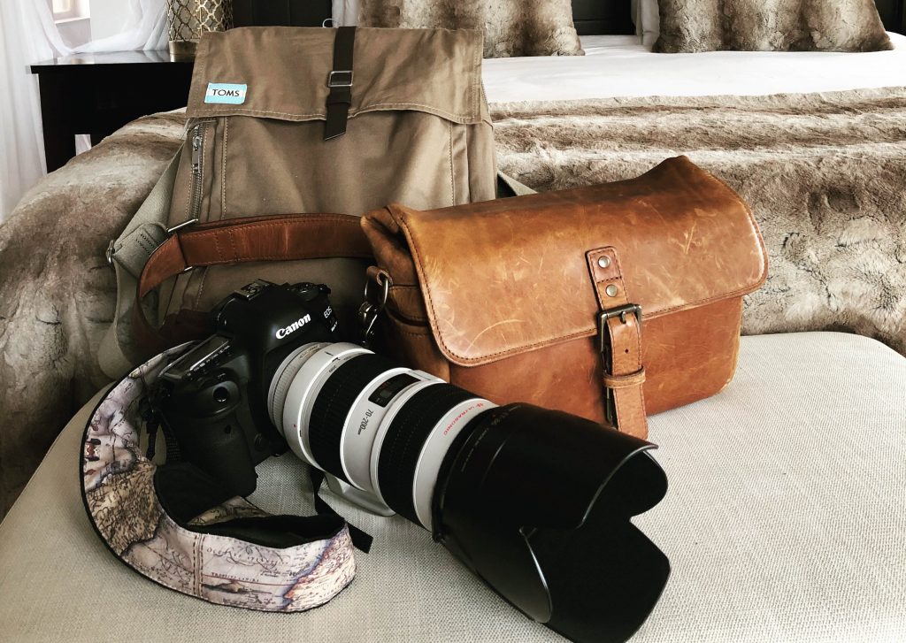 A camera, leather bag, and tan backpack sitting next to a bed. Learn what you need to bring on your next African adventure with this essential safari packing guide. And for more travel tips follow The Scenic Suitcase on Pinterest!
