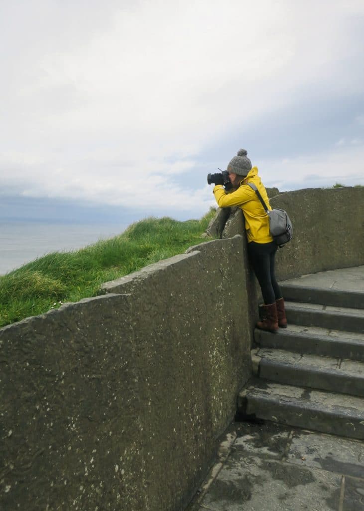 A girl in a yellow jacket taking pictures against a stone wall at the Cliffs of Moher.