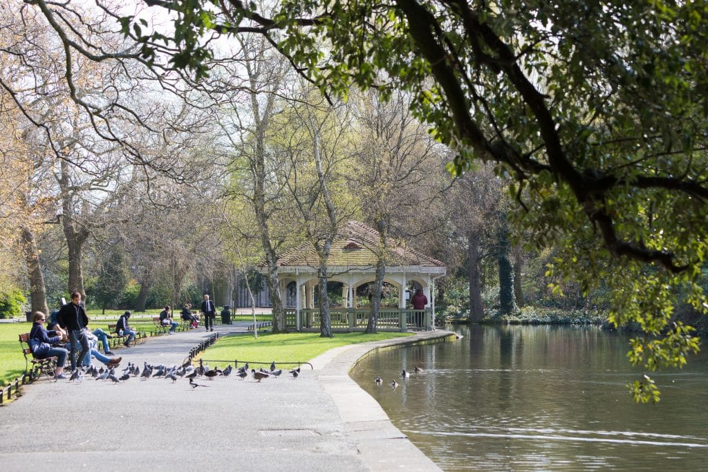 People feeding pigeons along a lake with a gazebo in the background. #StStephensGreen