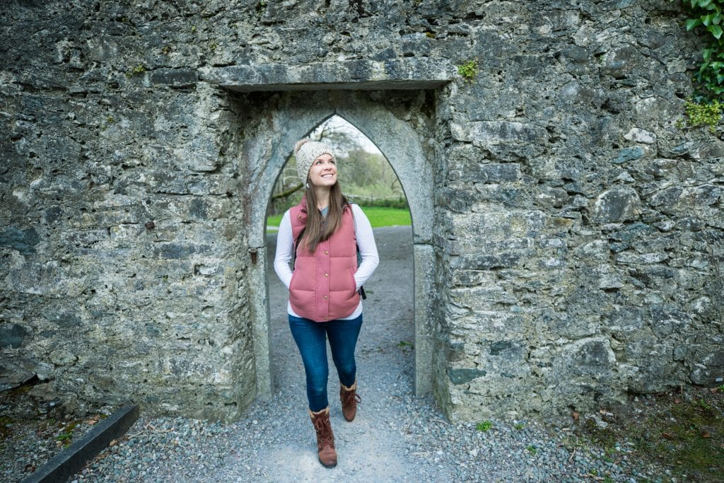 A girl wearing a pink down vest walking through a stone archway. #muckrossabbey