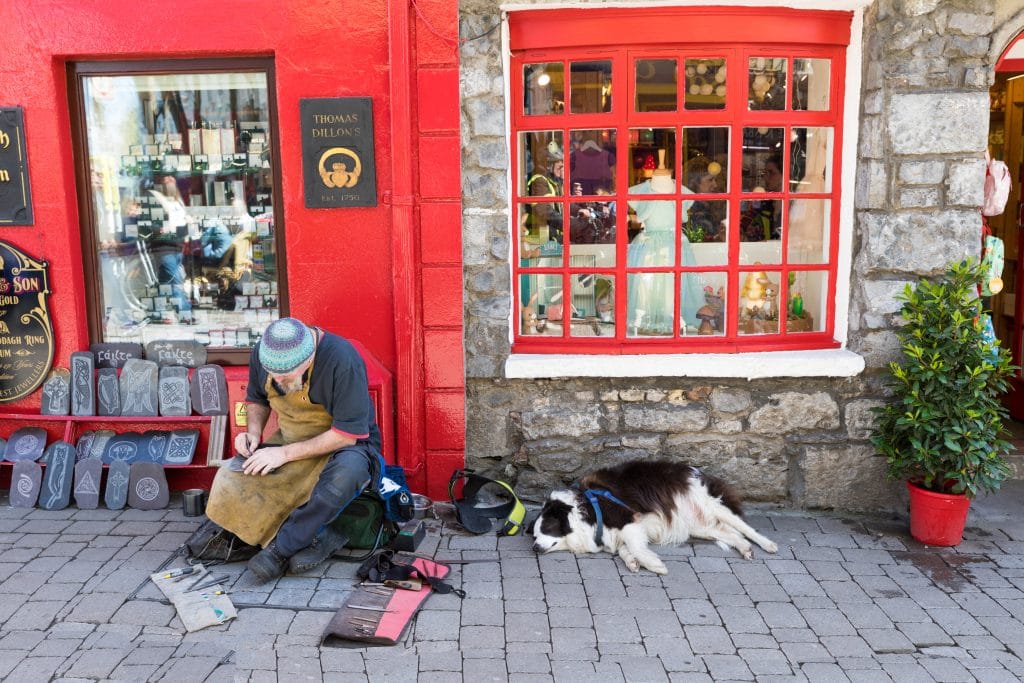 An artisan carving stones with his dog next to a red storefront in Galway. There are countless reasons to visit Ireland. Discover 8 of the most amazing places you really shouldn't miss on your next trip to the emerald isle!