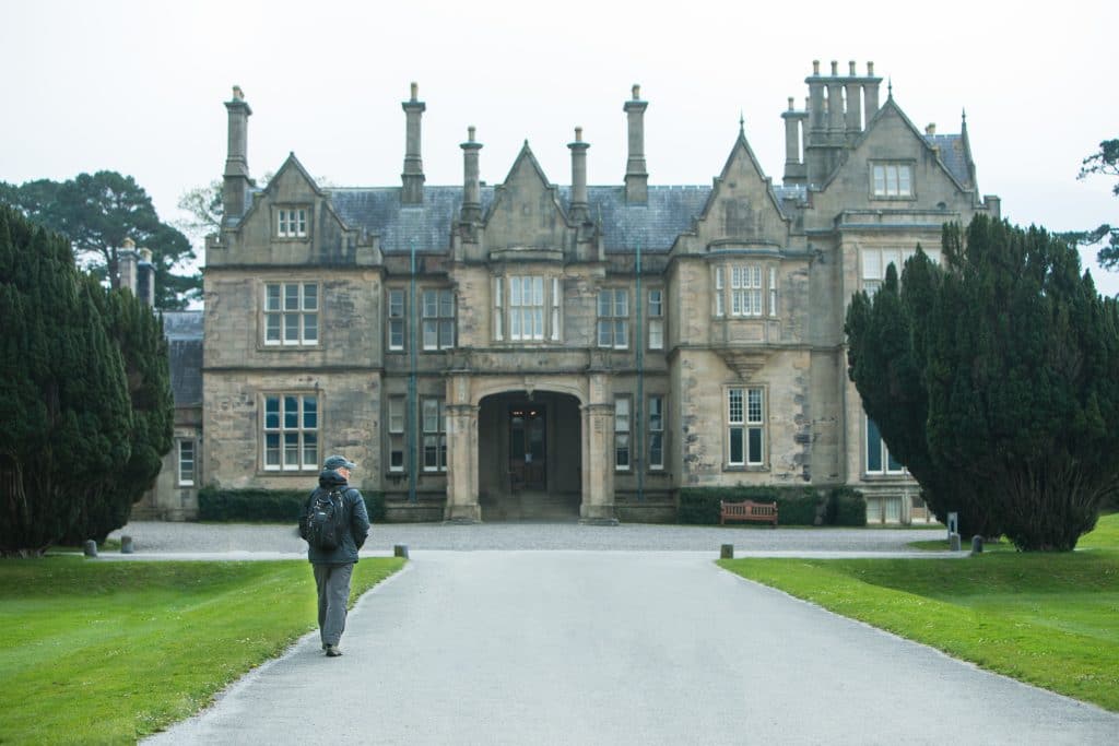The imposing fascade of the Muckross House with a man walking down the road towards it. It's one of the most amazing things to see in Killarney! #muckrosshouse #killarney