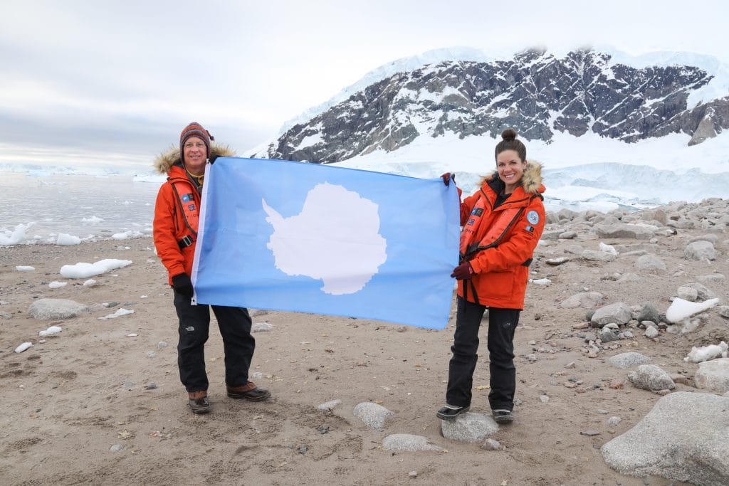 Two people holding an Antarctica flag on a beach with icebergs and a glacier. Discover the many reasons to visit Antarctica, and what to expect when you get there. From impressive icebergs to playful penguins and more! #antarctica
