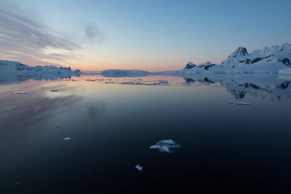 Sunset clouds and snowy mountains reflected in the water in Antarctica. Discover the many reasons to visit Antarctica, and what to expect when you get there. From impressive icebergs to playful penguins and more! #antarctica