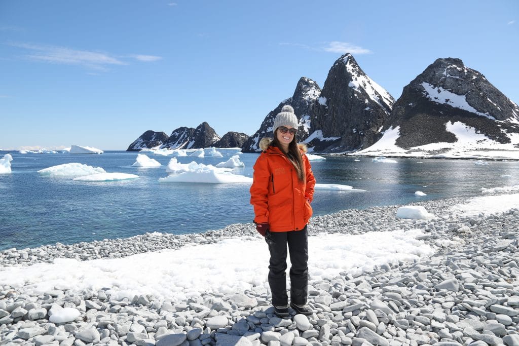 A girl in an orange jacket standing on the edge of a pebble beach with black mountains in the background. #antarctica