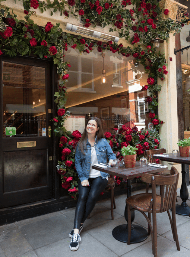 A girl sitting at a table next to a building covered in red flowers. #london