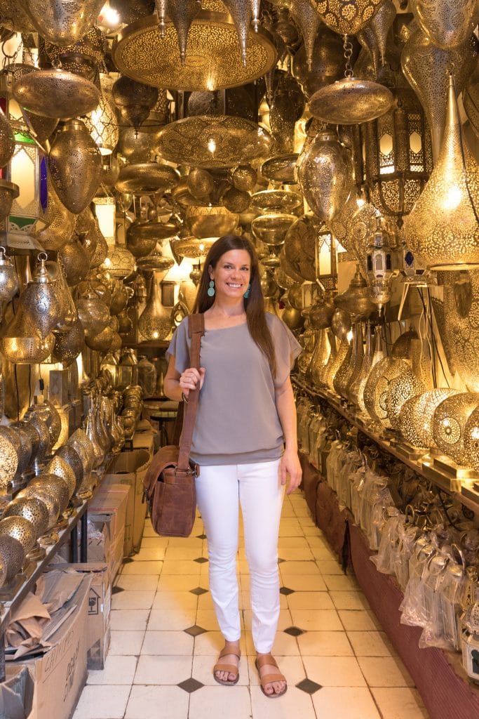 A girl in white pants and a grey shirt standing in a gold lamp shop in Marrakesh.