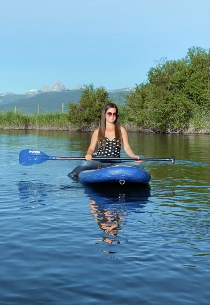A girl in a black and white polka dot bathing suit riding on a paddleboard down a river near the Grand Tetons.
