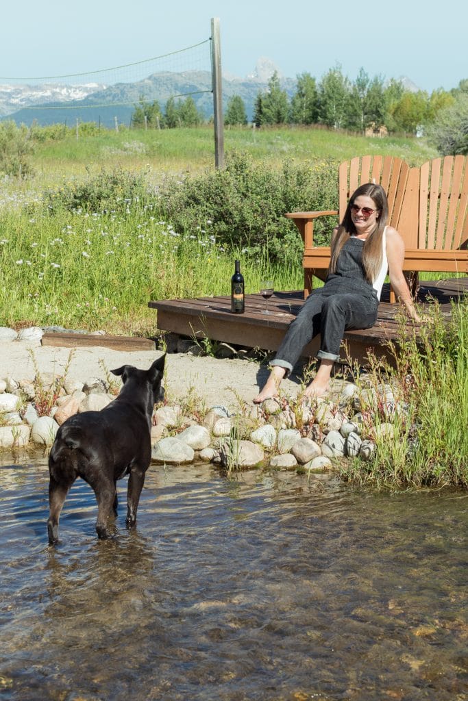 A girl sitting on a dock with her feet in the sand looking at a black dog standing in a creek.