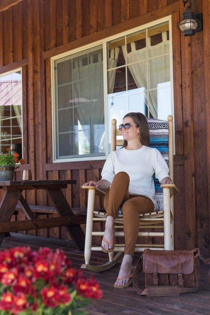 A girl sitting in a rocker on a cabin porch with red flowers in the foreground.