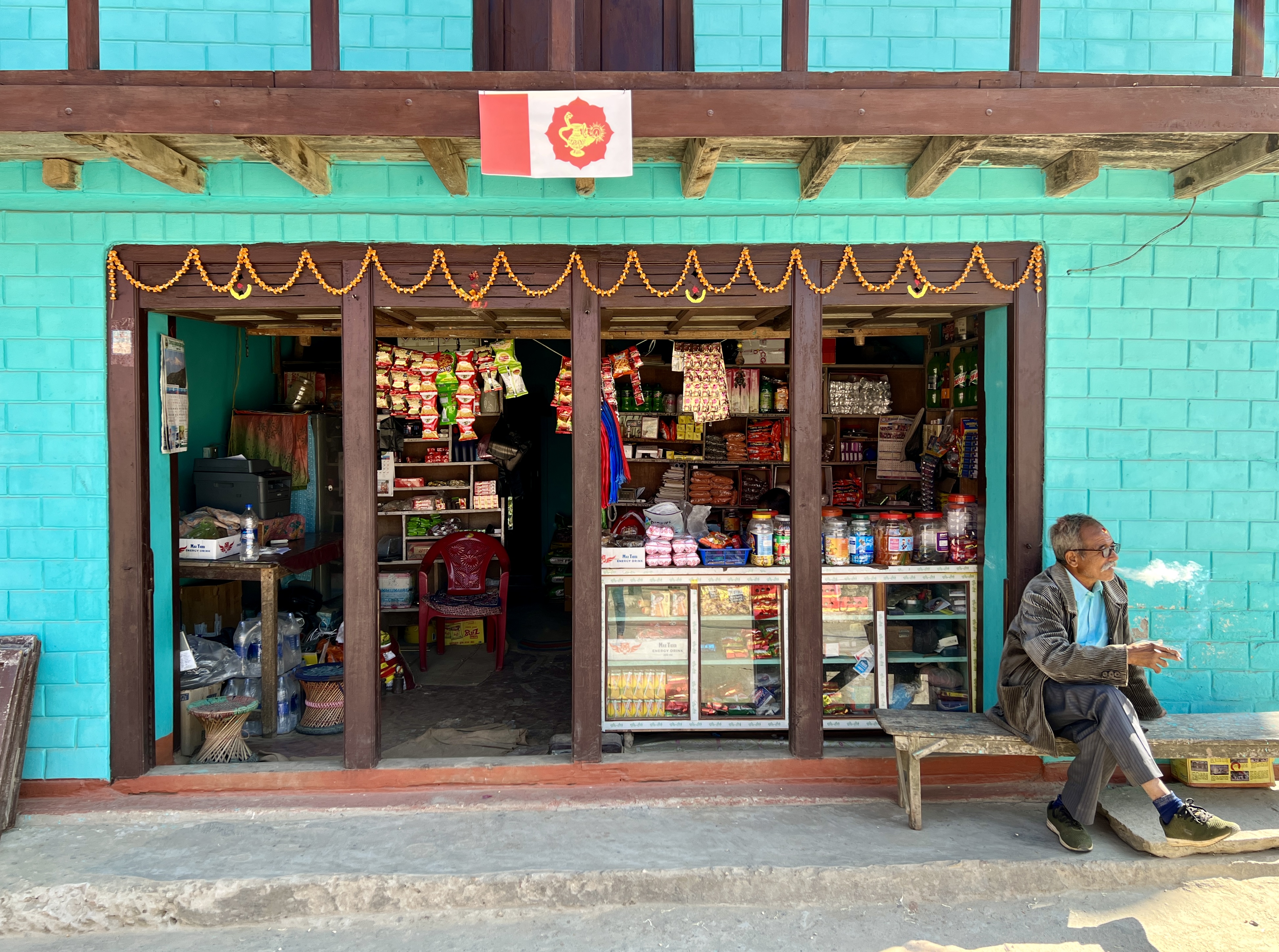 A man sitting and smoking next to a bright green store in Khandbari, Nepal.