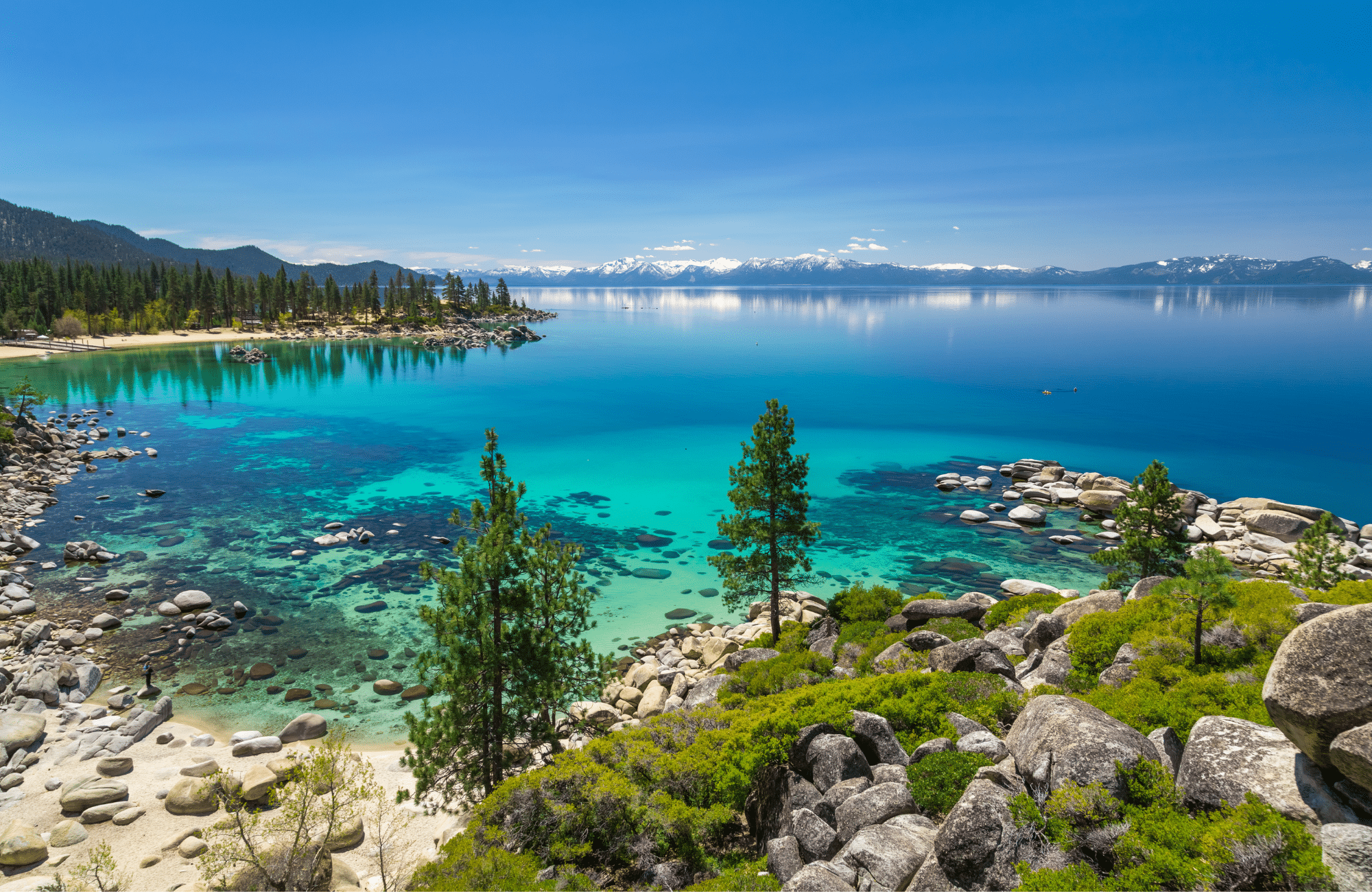 Large rocks and a sandy shoreline of a bright blue lake near mountains.
