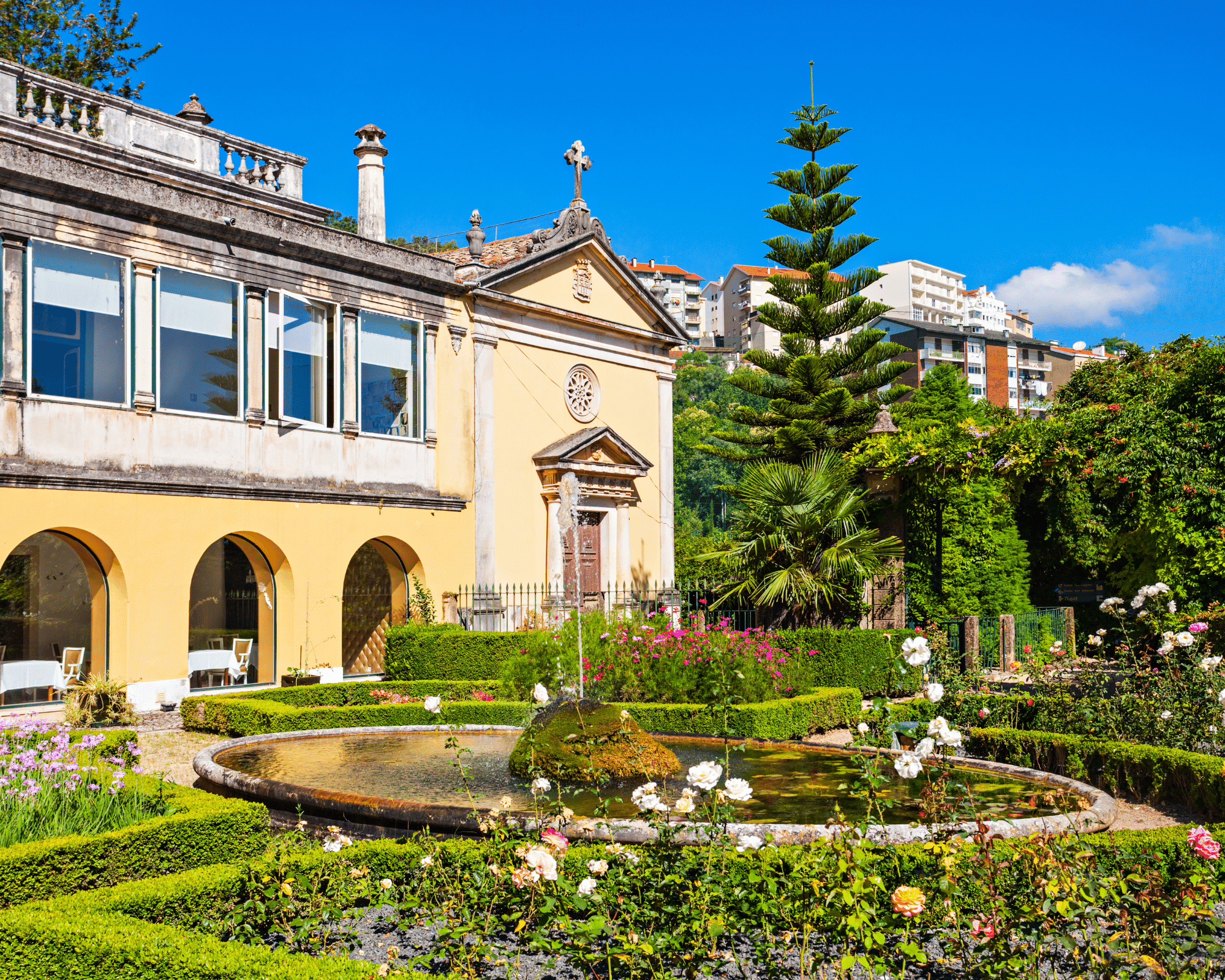 A cream colored building surrounded by a gardens with a fountain.